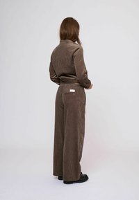 Barna kordbársony öltözet, enyhén testre szabott dzsekivel és bőszárú nadrággal, függőleges bordázattal és egy címkés hátsó zsebbel.