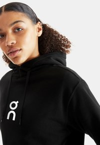 Juodas džemperis su kapišonu, pagamintas iš minkštos medžiagos, su kengūros kišene ir raišteliais. Priekinėje dalyje matomas baltas logotipas.