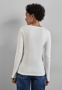 Weißes Langarmshirt mit rundem Halsausschnitt, aus weichem Baumwollmaterial; kombiniert mit blauen Jeans aus Denim mit Gesäßtaschen und subtiler Textur.