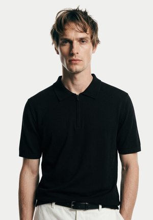 Junger Mann mit kurzen braunen Haaren, der ein schwarzes Poloshirt mit kurzen Ärmeln und Frontreißverschluss sowie weiße Hose vor einem schlichten Hintergrund trägt.