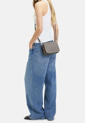 Nainen valkoisessa hihattomassa paidassa ja väljissä sinisissä farkuissa, jolla on pieni harmaa olkalaukku pitkällä hihnalla olkapään yli.