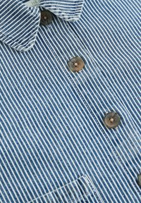 Blau-weiß gestreiftes Stoffhemd mit Kragen, ausgestattet mit zwei braunen Knöpfen und einer Brusttasche. Material aus Baumwolle, glatte Textur.
