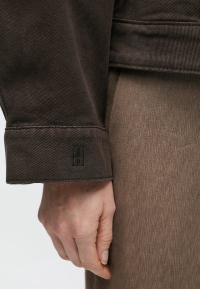 Rudos korduroinės striukės apykaklė su siuvinėtomis detalėmis ir ant rankogalių išsiuvinėtu logotipu. Matoma ranka, laikanti šviesiai rudą audinį.