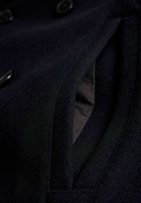 Jūras spēku krāsas vilnas mēteļa audums ar gludu tekstūru, ar slēptu kabatas atvēršanu un melniem pogām gar priekšpusi.