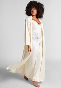 Vrouw met krullend haar, gekleed in een vloeiende crèmekleurige robe over een bloemenprint slipdress, die blootvoets loopt op een effen grijze achtergrond.