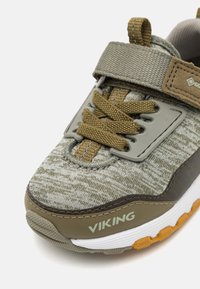 Grå og olivensneaker med teksturert stoff, elastiske lisser, justerbar stropp og gummisåle med oransje detaljer. "VIKING"-logoen er synlig.