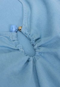 Tessuto azzurro chiaro con un design arricciato al centro, dotato di una piccola perla blu e un accento metallico dorato. Texture morbida.