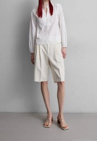 Valkoinen pitkähihaiste shirt, jossa on osittainen nappilista, yhdistettynä beigeihin pleatattuihin shortseihin ja nudeihin korkokenkiin, joissa on hihnat yksityiskohtina.