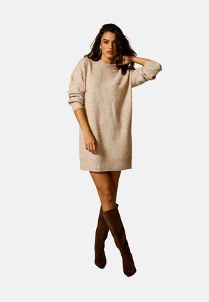 Robe en tricot de couleur beige clair avec un col et des poignets côtelés. Manches longues et coupe décontractée, assortie à des bottes brunes jusqu'aux genoux.