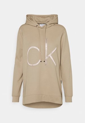 Beżowa bluza z długim rękawem, z regulowanym sznurkiem z przodu i dużym, błyszczącym logo "CK" na środku klatki piersiowej.