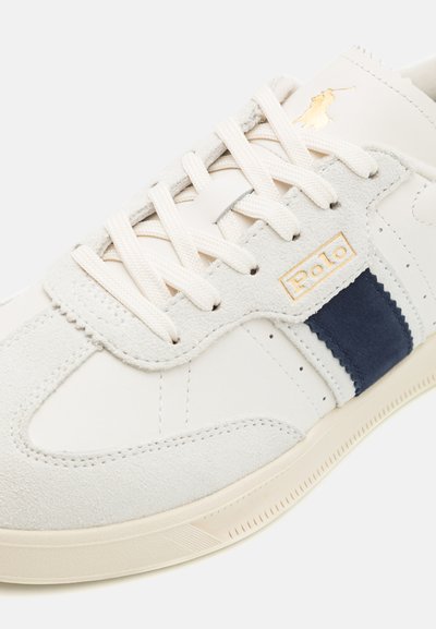 White suede sneaker with navy stripe and tan accents. Features laces, perforated detailing, and a gold "Polo" logo on the side.