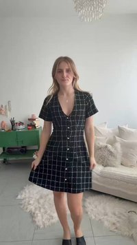 Vrouw staat binnen in een zwart geruit jurkje met knopen, houdt de zoom van de jurk vast, met een witte bank en een groene kast op de achtergrond.
