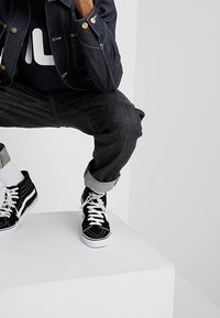Černé vysoké tenisky s bílými šňůrkami, spárované s tmavými džíny a tmavou bundou. Outfit zahrnuje manžety na nohavicích a tričko s grafickým potiskem.