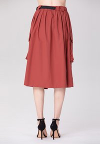Jupe longueur genou couleur rouille avec des plis, poches latérales et une taille élastique noire, associée à des chaussures à talons hauts noires.