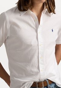 Vit kortärmad skjorta med strukturerad yta, med en klassisk krage och en blå broderad logotyp på bröstet. Knäppning fram.
