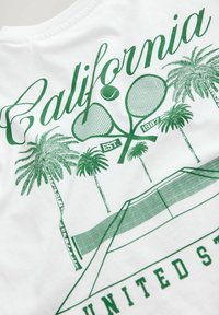 Witte t-shirt met groene grafische print van tennisrackets, palmbomen en de tekst "California EST. 1987," met een illustratie van een tennisbaan.