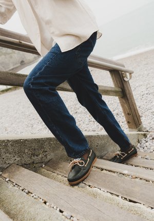 Person iført mørke jeans og sorte mokkasinsko klatrer på trætrapper ved en småstensstrand med et metal- og trægelænder.