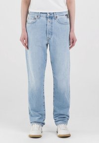 Person som bär ljusblå raka jeans, vita sneakers och en vit skjorta som står mot en enkel bakgrund.