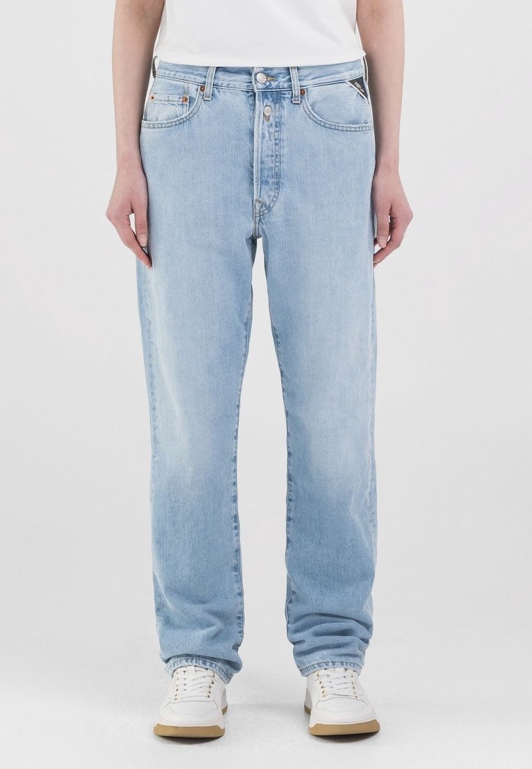 Person som bär ljusblå raka jeans, vita sneakers och en vit skjorta som står mot en enkel bakgrund.