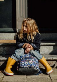 Blauwe rugzak met hondenpatronen, gewatteerd en met ritssluiting. Gele rubberen laarzen, zwarte jas en spijkerbroek, zittend op de trappen.