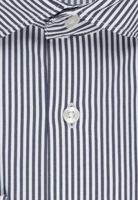 Bluse mit Knöpfen aus Baumwolle, präsentiert in Marine- und Weiß mit vertikalen Streifen und einem glatten Textur, versehen mit einem Knopf am Kragen.