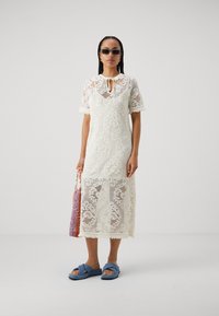 Robe en dentelle blanche à manches courtes, décolleté en keyhole et ourlet frangé. Associée à des sandales glissantes bleues et à un sac tissé multicolore.