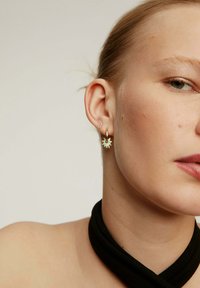 Pendientes de aro dorados con un diseño de estallido estelar, que cuentan con un acabado texturizado y pequeñas piedras de acento. Llevados por una modelo con una blusa negra sencilla.