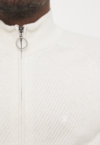 Světle krémový zip-up svetr vyrobený z texturované pleteniny. Má vysoký límec, stříbrný zip s kruhovým tahem a decentní výšivku loga.