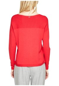 Pull tricot rouge avec un large décolleté, texture côtelée aux épaules et manches longues. Accompagné d'un pantalon gris clair.