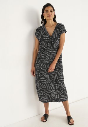Woman stands against a white wall, wearing a black dress with a leaf pattern and black slide sandals, smiling confidently.
