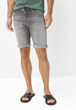 Mann trägt hochgekrempelte graue Jeansshorts, ein grünes T-Shirt und schwarze Ledersandalen, steht auf einer weißen Oberfläche.