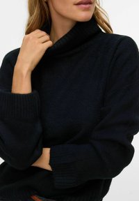 Tumši zils adīts džemperis ar augstu ribotu apvalka apkakli un platām piedurknēm, ar teksturētu audumu. Brīvs siluets ar smalku spīdumu.