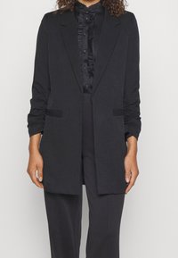 Černý přizpůsobený blazer s dlouhou siluetou, s nařasenými rukávy a dvěma předními kapsami. Nositý přes odpovídající blůzu s volánkovými detaily.