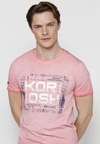 Rožinė medvilnės marškinėliai su apvalia iškirpte, kuriuose yra drąsus baltas ir tamsiai mėlynas grafikų dizainas su tekstu "KOROSH" ir papildomais akcentais.
