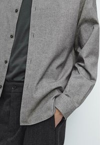 Camicia grigia con bottoni, in tessuto strutturato, maniche lunghe e vestibilità comoda. Indossata sopra una maglietta scura e abbinata a pantaloni scuri.