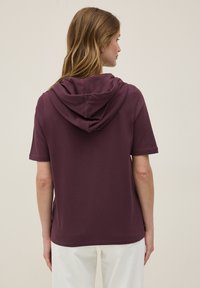Camiseta de capucha de color marrón con mangas cortas, hecha de una tela suave. Presenta un corte relajado y una costura en el hombro.