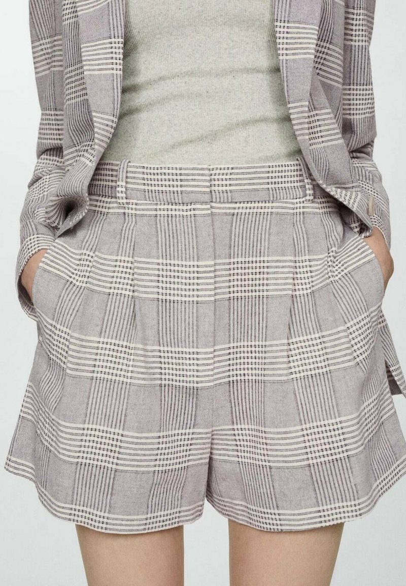 Femme portant un short à carreaux beige taille haute et un blazer assorti, les mains dans les poches, associé à un haut en maille beige clair.