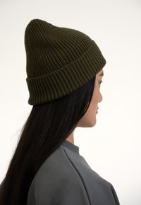 Caciuce de tricot de culoare verde măsliniu, cu vârful conic, textură côtelată și manșetă pliată, purtată de o persoană cu păr lung și negru, pe un fundal deschis.