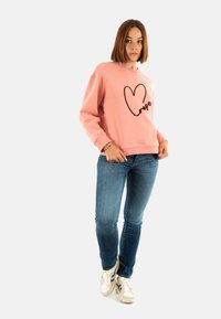 Sudadera rosa con un corazón negro y el texto "amor", combinada con vaqueros azules y zapatillas blancas. Diseño casual, textura suave, corte relajado.