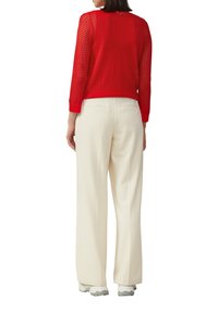 Roter, offener Strickpullover mit Rundhalsausschnitt und langen Ärmeln, kombiniert mit cremefarbenen Hosen mit Gesäßtaschen und entspannter Passform.