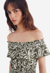 Grünes schulterfreies Kleid mit weißen Blattmustern, Rüschenausschnitt und strukturierter Stoff. Verstellbarer elastischer Taillenbund und Pom-Pom-Akzente.