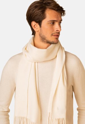 Junger Mann mit kurzem braunem Haar trägt einen cremefarbenen Schal und einen passenden Pullover und schaut nach rechts vor einem schlichten hellen Hintergrund.