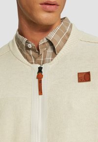 Cremefarbenes Strickpullover mit Reißverschluss, geripptem Kragen und braunem Leder-Logo-Patch. Darunter ist ein braun kariertes Hemd sichtbar.
