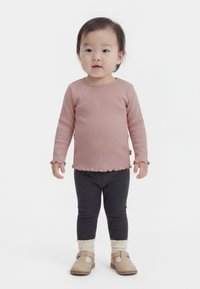 Kisgyermek áll, hosszú ujjú, poros rózsaszín felsőt, sötétszürke leggingset, krémszínű zoknit és bézs cipőt visel, fehér háttér előtt.