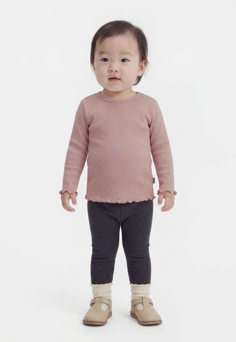 Kisgyermek áll, hosszú ujjú, poros rózsaszín felsőt, sötétszürke leggingset, krémszínű zoknit és bézs cipőt visel, fehér háttér előtt.