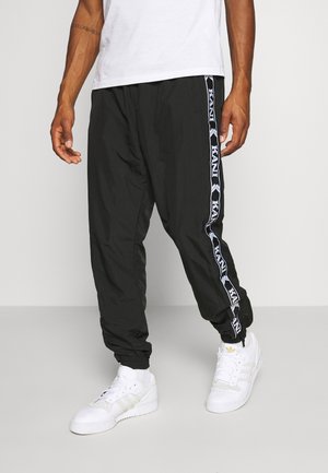 Mann trägt schwarze Jogginghose mit weißen Logostreifen an den Seiten und weiße Sneakers, steht vor einem einfarbigen Hintergrund.