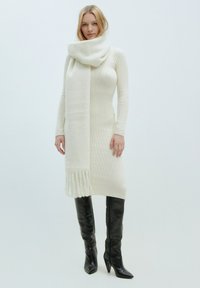 Strickkleid in creme mit Rippstrickmuster und langem, weißen Schal mit fransigen Kanten. Das Modell trägt schwarze, spitz zulaufende Stiefel bis zum Knie.