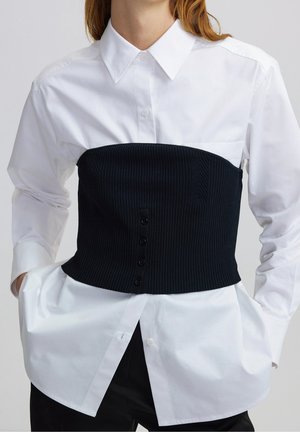 Frau trägt ein weißes, langärmliges Hemd mit Knöpfen und darüber ein schwarzes, geripptes, trägerloses Korsett-Top.