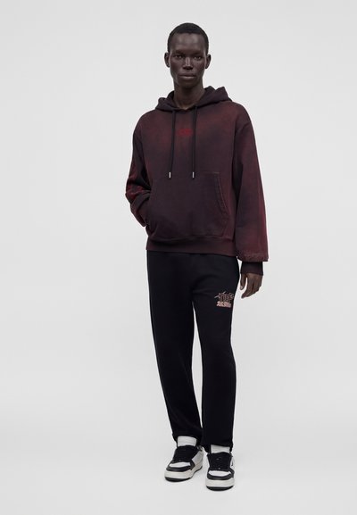 Jeune homme portant un sweat à capuche rouge foncé et un pantalon noir avec des baskets noires et blanches, debout devant un fond clair uni.