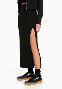 Black maxi skirt with high side slit, made from stretch fabric. Paired with black sneakers featuring gum soles and white accents.